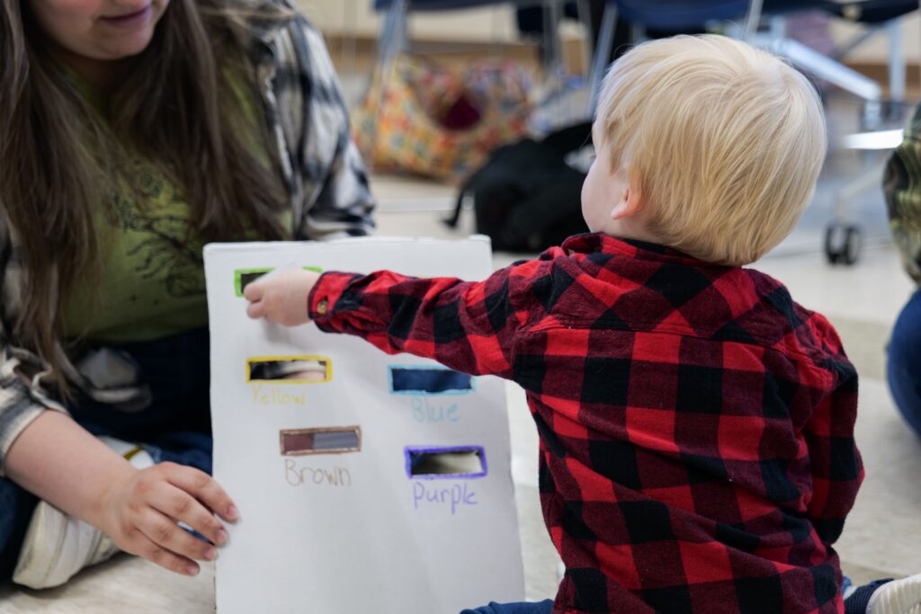 A young child with blonde hair wearing a red and black plaid shirt points to a color swatch on a white board during a learning activity. The board displays several color samples labeled “Yellow,” “Blue,” “Brown,” and “Purple.” An adult next to the child, partially visible, holds the board steady and watches the interaction.