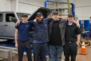 Four Eastern Maine Community College automotive students stand arm in arm in a campus auto lab, smiling in front of a gray Jeep with its hood open. From left: Daniel Tomoiu, Haris Plavsic, Nathaniel Kirkendall, and Gavin McDonough. All wear Mopar CAP hats and safety glasses, representing EMCC in the national Mopar Career Automotive Program Bracket Challenge.
