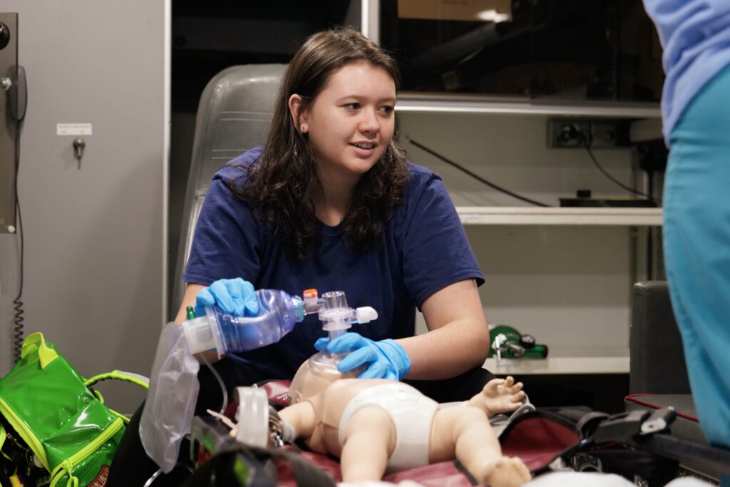 A student wearing blue medical gloves uses a bag-valve mask to provide ventilation to an infant training manikin during a hands-on emergency medical training exercise. She is seated in a simulated clinical or ambulance environment, focused on the procedure while another person stands nearby.