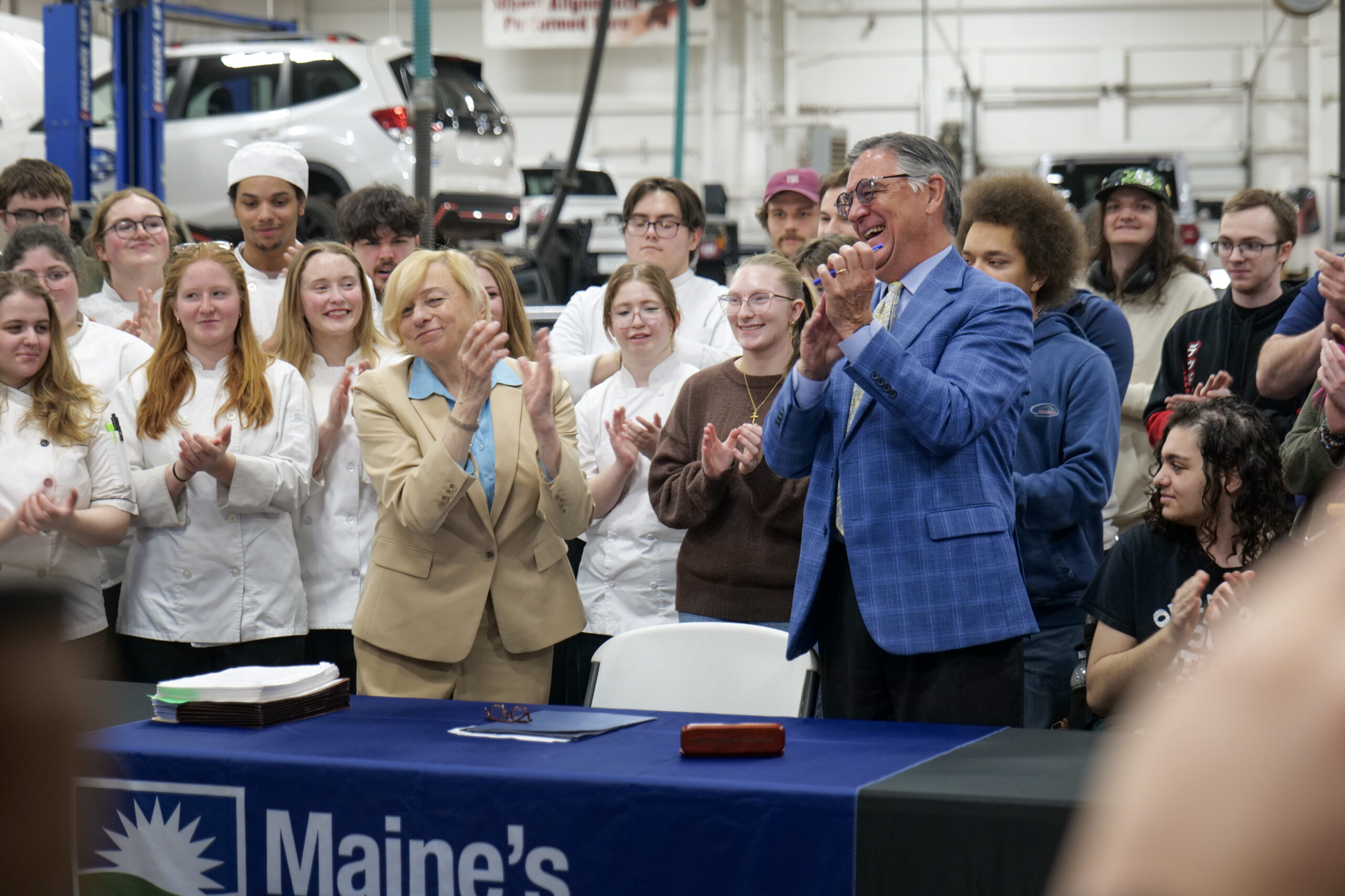 Governor and college leaders applaud as students gather around a table after a bill signing event inside an automotive lab.